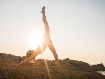 Side view of woman doing exercise on rock against sky during sunset