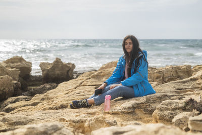 Young woman sitting on rock at beach against sky