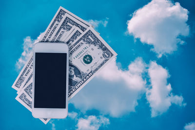 Digital composite image of mobile phone with paper currencies against cloudy sky