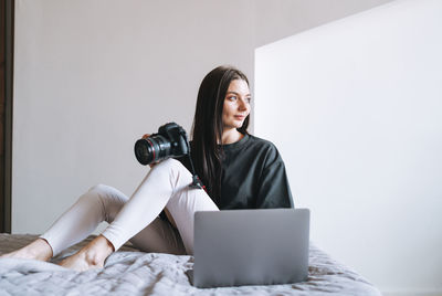 Young woman photographer with digital camera freelancer in casual using laptop on bed at home