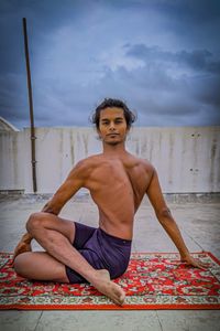 Portrait of shirtless man exercising on mat against cloudy sky