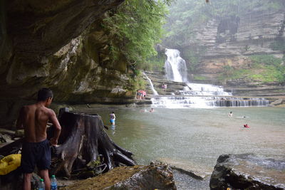 Shirtless man standing under cliff against waterfall