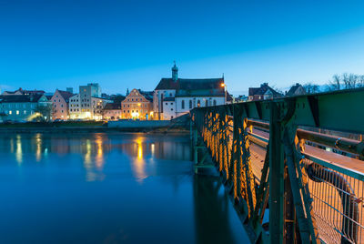 Illuminated bridge over river by buildings against sky at dusk
