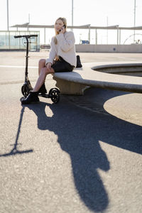 Young woman on the phone sitting on bench at sunlight with kick scooter