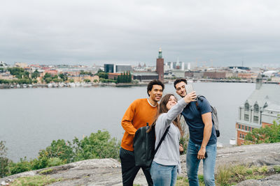 Smiling friends taking selfie with mobile phone while standing by river in city against sky