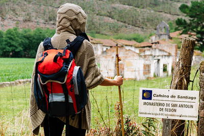 Back view of unrecognizable hiker with backpack and trekking stick standing against ancient monastery of san antolin de bedon during pilgrimage on camino de santiago in spain