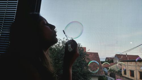 Side view of woman blowing bubble while standing by window at home