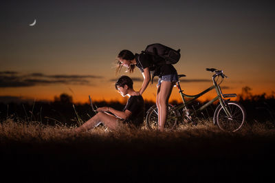 Side view of woman looking at man using laptop on land against sky during sunset