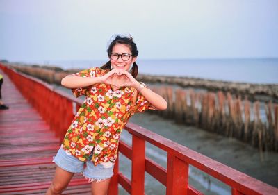 Portrait of smiling woman making heart shape on bridge against sky