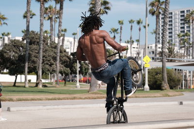 Rear view of shirtless man with bicycle against trees in city