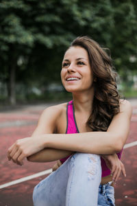 Smiling thoughtful young woman sitting against trees at park