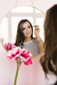 Happy beautiful young woman paints her eyelashes in front of the mirror at home