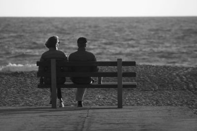 Rear view of couple sitting on bench at beach against sea