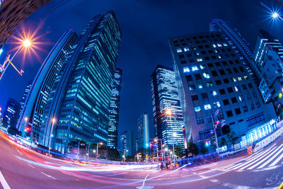 Light trails on city street by buildings against sky at night