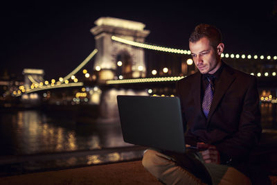 Businessman working on laptop by chain bridge at night