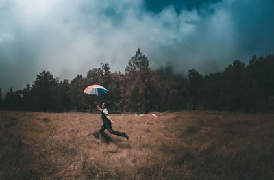 Side view of young man with umbrella running on grassy field against cloudy sky