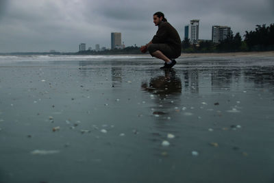 Man crouching at beach against sky at dusk