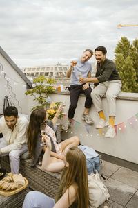 Smiling men toasting drinks with friends while sitting on wall in balcony