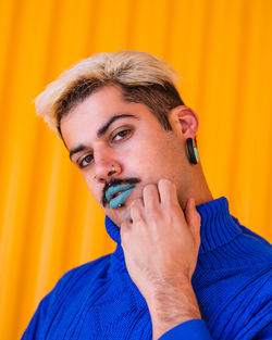Androgynous male with blue lips and in stylish wear standing on background of yellow wall in city while looking at camera and touching face