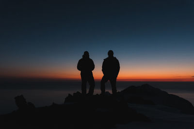 Silhouette men standing on rock at beach against sky during sunset