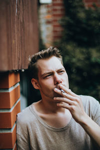 Young man smoking cigarette while leaning on brick wall