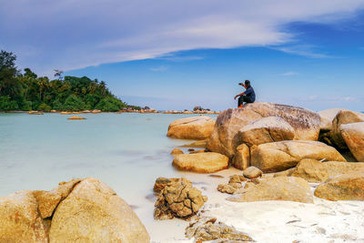 Man sitting on rock at beach against sky