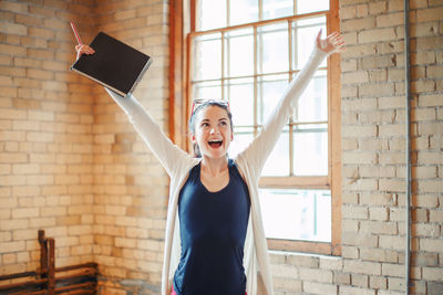 Cheerful woman with arms raised standing against brick wall