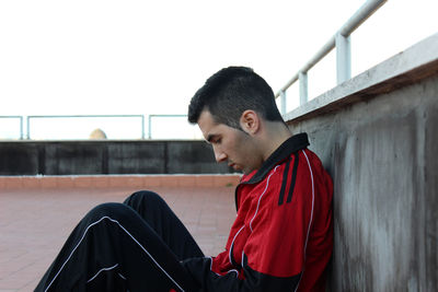 Side view of young man looking away while sitting on railing against wall