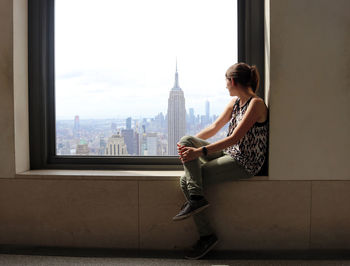 Woman sitting on window sill while looking at cityscape