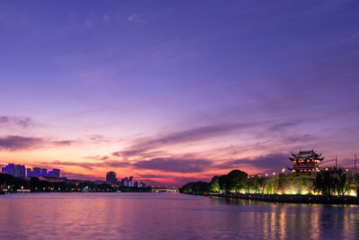 Illuminated buildings by river against sky during sunset