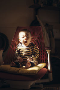 Portrait of cute baby boy yawning while sitting on chair at home