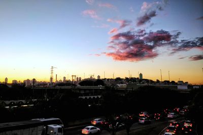 Cars on road by buildings against sky during sunset