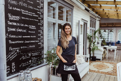 Portrait of smiling female owner with hands in pockets standing in restaurant