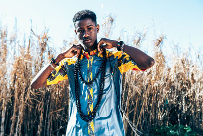 Front view of serious ethnic man wearing native african clothing and accessories and looking at camera while standing on field on sunny summer day