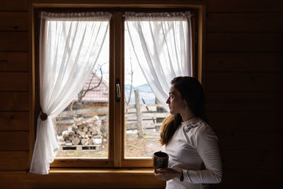 Woman sitting at the window of a mountain cabin, enjoying a cup of tea and the view.