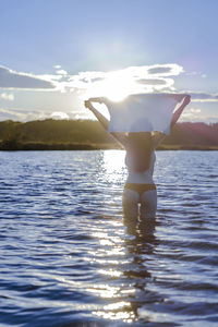 Rear view of woman with arms raised holding top standing in lake against sky