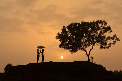 Silhouette couple with umbrella standing on hill against orange sky during sunset