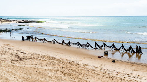 Several fishermen are seen walking in a line and carrying a large fishing net. 