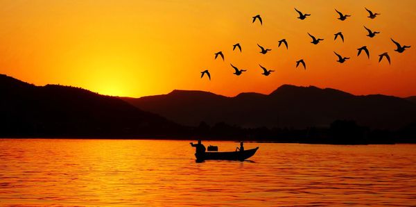 Silhouette people in boat on lake against orange sky during sunset