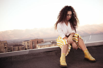 Young woman with curly hair sitting outdoors against sky