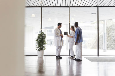 Full length of nurses and doctor discussing over digital tablet against window in corridor at hospital