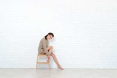 Full length portrait of woman sitting on stool against brick wall