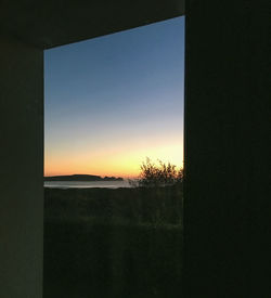 Silhouette trees on field against sky during sunset seen through window