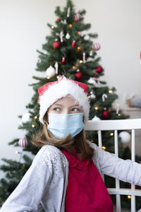 Cute girl wearing mask looking away while sitting against christmas tree at home