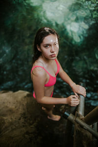 From above side view of young female traveler in pink bikini looking at camera while standing near railing on rocky shore of natural pool in rincon de la vieja national park in costa rica