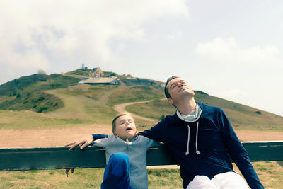 Father and son enjoying in sunlight with eyes closed while sitting on a bench at mountain peak.