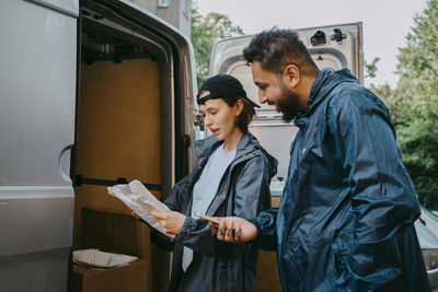 Male and female delivery coworkers reading details on package while standing near van