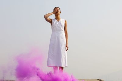Young woman covering eyes while standing by pink distress flare against clear sky
