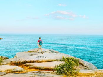 Rear view of man standing on cliff by sea against sky