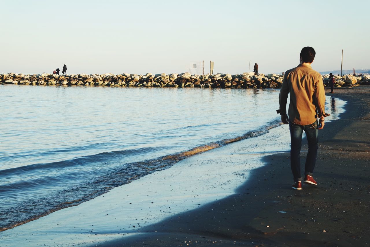 water, sea, clear sky, rear view, standing, lifestyles, full length, leisure activity, men, beach, copy space, shore, walking, person, casual clothing, sky, built structure, nature